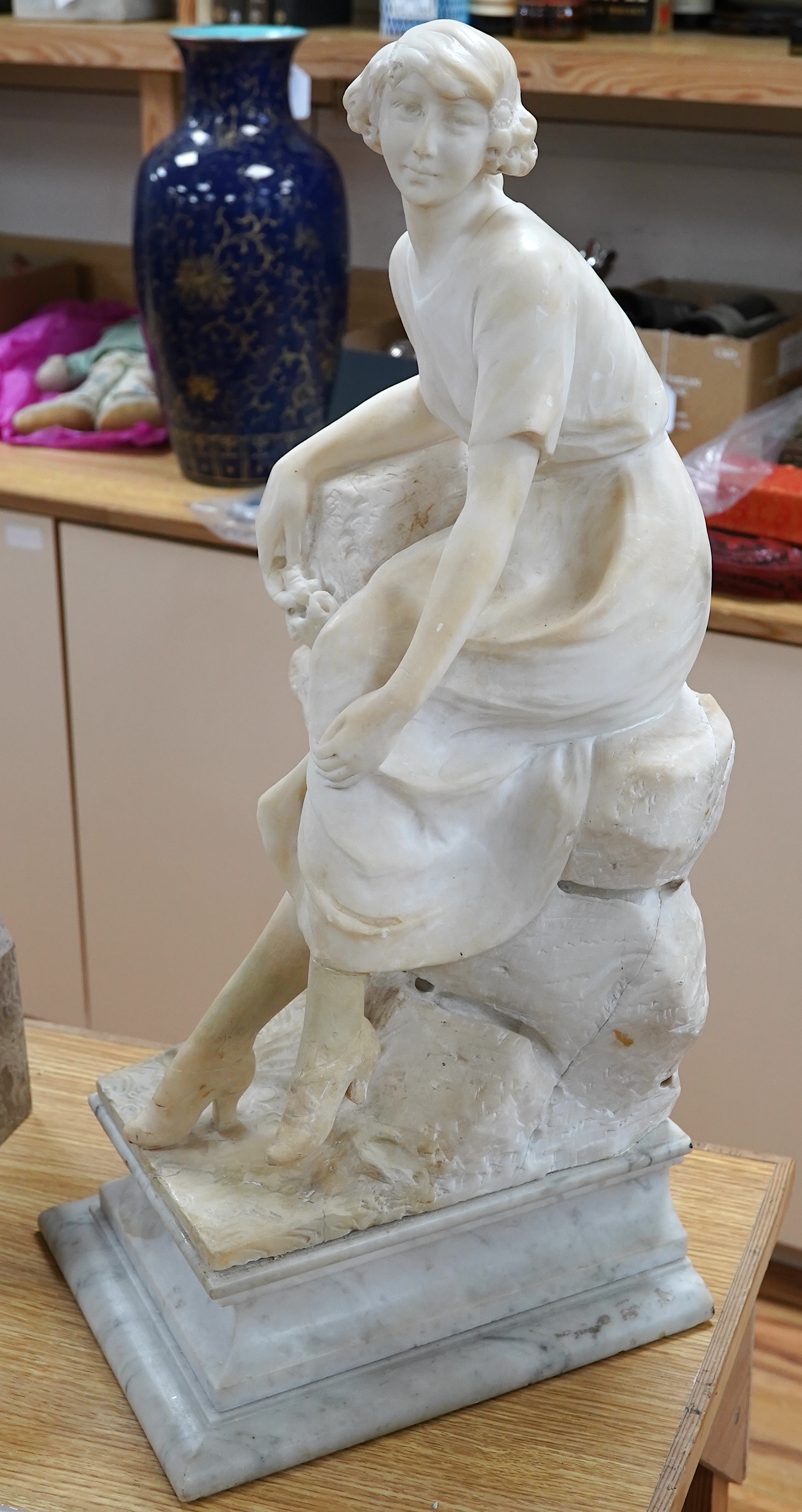 A French 1920’s-30’s alabaster sculpture of a lady seated on a stone wall, sculpture mounted on a marble stand, 63cm high. Condition - crack to alabaster at the front and discoloured restoration